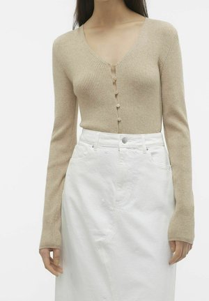 Femme portant un cardigan côtelé beige à manches longues boutonné, rentré dans une jupe blanche taille haute avec une broderie discrète en forme d'étoile.