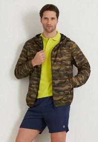 Kamouflagejacka i nyanser av grönt och brunt, med huva och elastiska ärmslut, parat med en ljusgul polo och marinblå shorts.