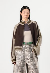 Brown and beige knitted bomber jacket with ribbed collar, featuring striped details and a zip front, worn over a fitted top and patterned pants.