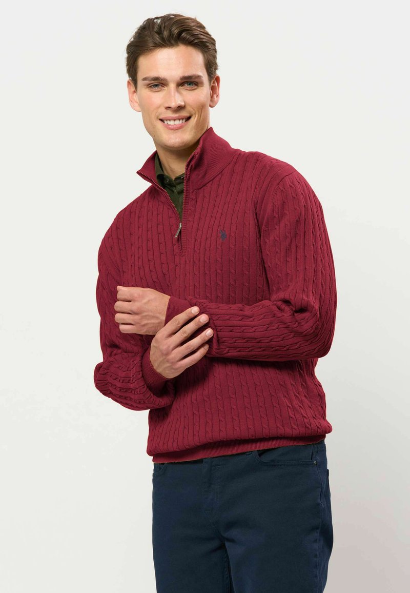 Jeune homme souriant portant un pull rouge en maille torsadée avec une fermeture éclair mi-hauteur et un pantalon bleu foncé, debout devant un fond clair uni.