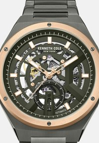 Het automatische horloge van Kenneth Cole heeft een groene wijzerplaat met gouden accenten, een skeleton ontwerp en een grijze armband van roestvrij staal.
