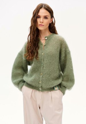 Kobieta z długimi falującymi włosami, ubrana w miękki zielony kardigan zapinany na guziki oraz jasnobeżowe spodnie z wysokim stanem na jednolitym tle.