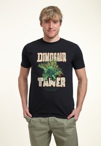 Joven vestido con una camiseta negra que tiene un estegosaurio verde y las palabras "Domador de Dinosaurios" en letras grandes.