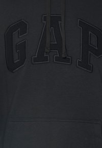 Svart hoodie med upphöjda texturerade bokstäver "GAP" över bröstet och en framficka i kängurustil med snörning vid halsen.