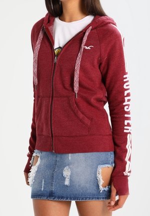 Kobieta ubrana w czerwoną bluzę z kapturem na zamek z napisem "Hollister" na rękawie i zniszczoną niebieską mini spódniczkę jeansową.