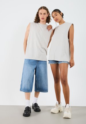 Helles graue ärmelloses Shirt kombiniert mit einem Mann in blauen Jeansshorts und schwarzen Slippern sowie einer Frau in Jeansshorts und weißen Sneakers.