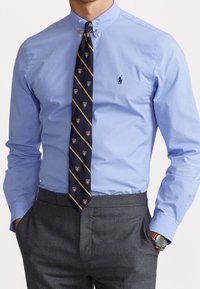 Man som bär en ljusblå skjorta med knappar, marinblå randig slips med sköld, grå byxor och en klocka med brunt läderarmband, med händerna i fickorna.