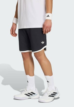 Osoba nosząca białe opaski na nadgarstki Adidas, czarne spodenki Adidas, białe skarpetki Adidas oraz białe sneakersy Adidas z czarnymi paskami.