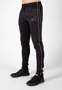Zwarte sportieve broeken van glad stof, met een gouden zijstreep en een ritssluitingzak. Gecombineerd met zwarte sportieve schoenen.