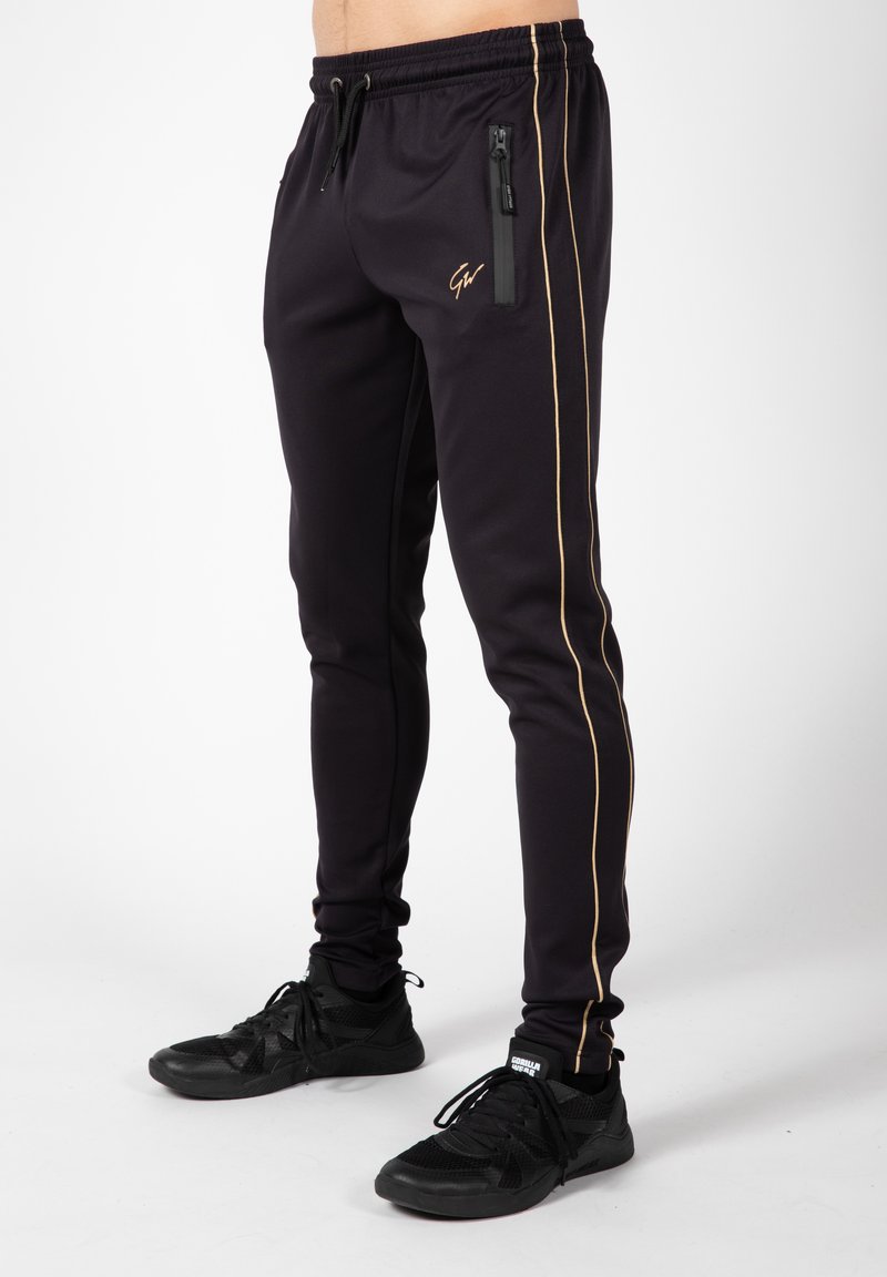 Zwarte sportieve broeken van glad stof, met een gouden zijstreep en een ritssluitingzak. Gecombineerd met zwarte sportieve schoenen.