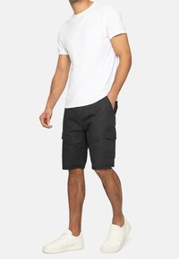 Mężczyzna w białych sneakersach, czarnych szortach cargo i gładkiej, białej koszulce z krótkim rękawem, stojący na jasnym tle.