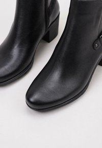 Bottes noires en cuir à la cheville avec un bout pointu, un talon épais, une fermeture éclair sur le côté et une texture lisse. Présente un petit accent dans la zone de la fermeture éclair.