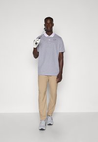 Striped polo shirt in white and navy, beige pants, holding a golf club, wearing a white golf glove, paired with gray sneakers.