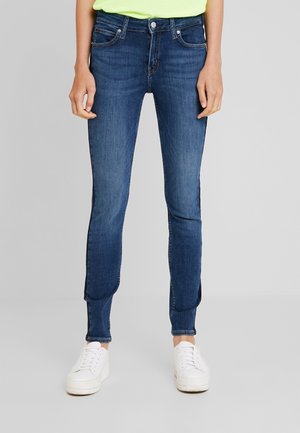 Slim fit donkerblauwe spijkerbroek gedragen met witte sneakers en een lichtgeel topje, getoond van taille tot voeten tegen een effen achtergrond.