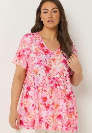 Kvinde iført en kortærmet V-hals top med et pink og rødt blomsterprint, stående foran en neutral baggrund.