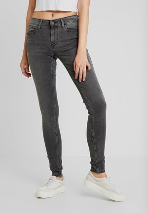 Kvinde iført højtaljede, vaskede sorte skinny jeans med hvide plateau-sneakers og en cropped hvid ribbet top, stående foran en ensfarvet grå baggrund.