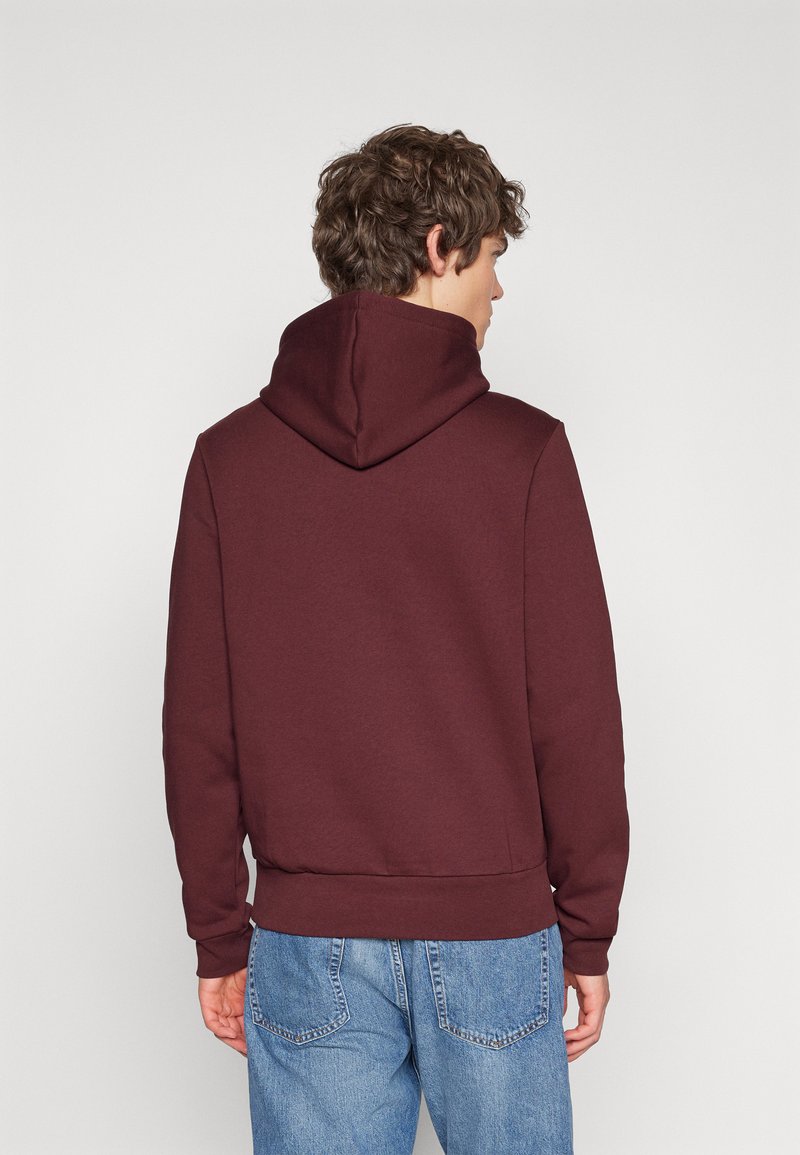 Bordeauxfarbener Hoodie mit strukturierter Oberfläche, ausgestattet mit einer großen Kapuze, gerippten Bündchen und einem Bund. Die Rückseite ist schlicht und hat keine sichtbaren Logos.