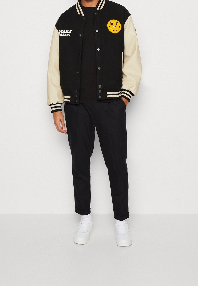 Homme portant une veste varsity noire avec des manches crème, texte "Tommy Jeans", un patch smiley jaune de NY, pantalon noir et baskets blanches.
