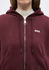 Personne portant un sweat à capuche zippé bordeaux avec le logo BOSS sur la poitrine et un pull crème à col montant en dessous, visible des épaules jusqu'à la bouche.