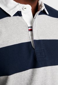 Gros plan d’un maillot de rugby rayé bleu marine et gris avec un col blanc et un détail rouge, blanc et bleu sur la patte de boutonnage.