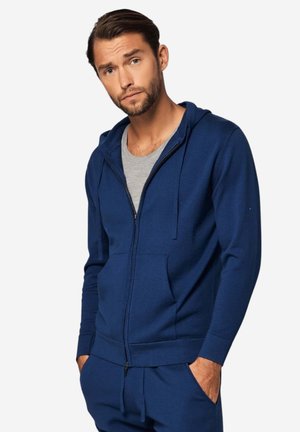 Mann mit kurzen Haaren und Bart, trägt einen marineblauen Kapuzenpullover mit Reißverschluss über einem grauen Shirt, Hände in den Taschen, vor einem einfarbigen Hintergrund.