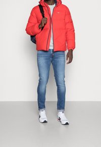 Rød polstret jakke med hætte, grå t-shirt, blå jeans og hvide sportsko med marineblå og røde accenter.