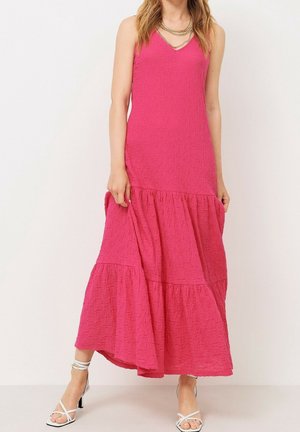 Kvinde iført ærmeløs, lyserød, tekstureret maxikjole med lagdelt skørt og hvide højhælede sandaler med stropper.