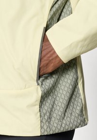 Žlutá bunda s texturovanou šedou podšívkou. Kapsa je částečně otevřená, odhalující vnitřní vzor a detaily zipu. Hladký vnější materiál.