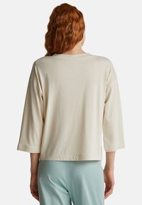 Frau mit langen roten Haaren, die ein lockeres beige Langarmoberteil und helle blaue Hosen trägt, mit dem Rücken zu einer schlichten weißen Wand.