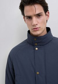 Tummansininen vuorattu takki korkealla kauluksella, jossa on kultaiset nepparit ja sivutasku. Sileä textura, moderni muotoilu.