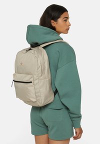 Mochila bege clara com alças acolchoadas, bolso frontal com fecho e logótipo. Usada sobre um moletom verde, com um corte descontraído e calções.