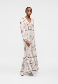 Femme debout portant une longue robe blanche brodée de fleurs, à manches longues et col en V, sur un fond uni.