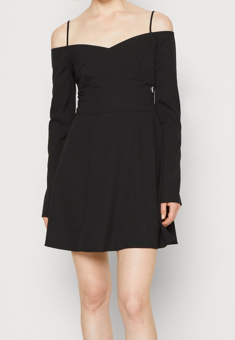 Svart klänning med långa ärmar, av-axel-design, v-ringning, figursydd midja, utsvängd kjol och silverdetaljer vid midjan.