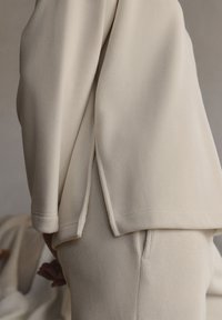 Pullover beige a coste con spacchi laterali, indossato sopra pantaloni coordinati a coste. Il tessuto appare morbido con una finitura testurizzata.