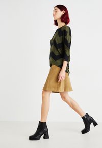 Suéter con patrón de zigzag en verde y negro, falda de pana color beige y botines negros de tacón. La modelo posa de perfil contra un fondo plano.