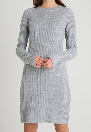 Neulottu vaaleanharmaa pitkähihaainen ribbineulottu villapuku, jota henkilö käyttää kädet ristittyinä vyötäröllä.