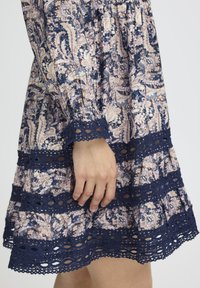 Jūras spēku ziedu kleita ar paisley rakstu, teksturētu mežģīņu apdari un A-līnijas siluetu, ar pūkainām piedurknēm un savilktu jostasvietu.