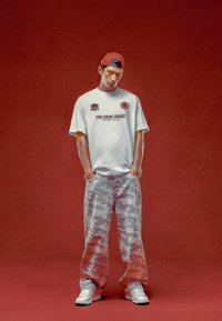 Siva grafička majica s natpisom "THE RHINO BRAND", uparena s laganim širokim hlačama s uzorkom kamuflaže i bijelim tenisicama. Crvena pozadina.