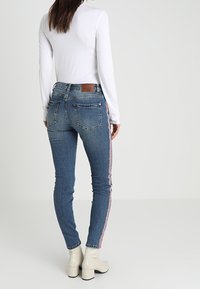 Smala blå denimjeans med röda och vita sidränder, med en midjehöjd som är mitt emellan och bakfickor. Byxorna är kombinerade med en långärmad vit topp.