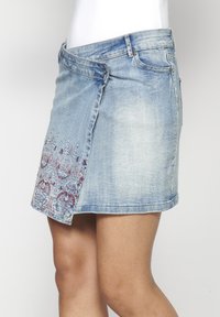 Denim krilo v svetlo modri barvi z bočno ploščo, ki vsebuje zapleteno cvetlično vezenje v rdeči in modri barvi. Standardna zasnova pasu, dolžina do sredine stegna.