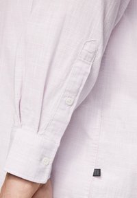 Lichtroze, langekleurenshirt met een gestructureerd, ruitpatroon; heeft witte knopen en een klein zwart merklabel bij de zoom.