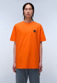 Lys orange bomulds-t-shirt med korte ærmer, rund hals og et lille cirkulært patch på venstre bryst. Modellen har grå bukser på.