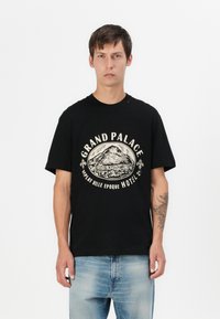 T-shirt nera in cotone con una grafica rotonda raffigurante una montagna, testo a bordo "GRAND PALACE" e una vestibilità rilassata, abbinata a jeans azzurri chiari.