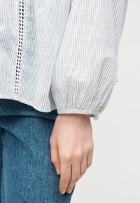 Χέρι ακουμπισμένο στο πλευρό φορώντας λευκό μπλουζάκι με μπλε ρίγες και μπλε τζιν παντελόνι.