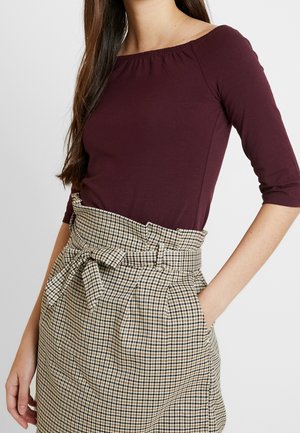 Kvinde iført en bourgognefarvet bluse med trekvartlange ærmer og en beige ternet nederdel med høj talje og bindebånd, hånd i nederdelslommen.