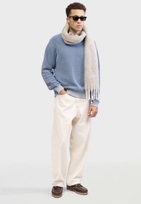 Světle modrý žebrovaný svetr, krémové kalhoty s širokými nohavicemi a béžová texturovaná šála s třásněmi. Oblečení doplňují hnědé boty. Nohušky jsou nasazené.