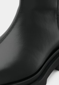 Μαύρο δερμάτινο μποτάκι αστραγάλου με λεία υφή, στρογγυλεμένη μύτη και χοντρή σόλα. Λεπτομέρεια ραφής κατά μήκος της άκρης προσθέτει διακριτικά σχεδιαστικά στοιχεία.