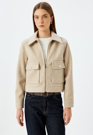 Beige kurz geschnittene Jacke mit großen Vordertaschen, einem Reißverschluss vorne und einem spitzen Kragen. Aus strukturiertem Stoff gefertigt, kombiniert mit dunklen Jeans.
