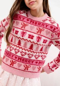 Fată îmbrăcată într-un pulover roz de Crăciun decorat cu inimioare roșii și roz, fundițe, brazi, cadouri și cuvântul "MERRY" în modele festive.