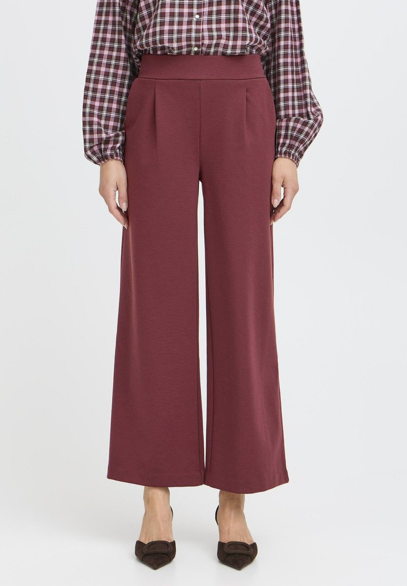 Pantaloni largi dintr-un material burgundy închis, cu pliuri frontale, talie înaltă și o textură netedă. Asortați cu tocuri întunecate, rotunde.